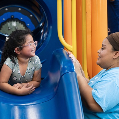 Child smiling on a playground slide talking with a caregiver