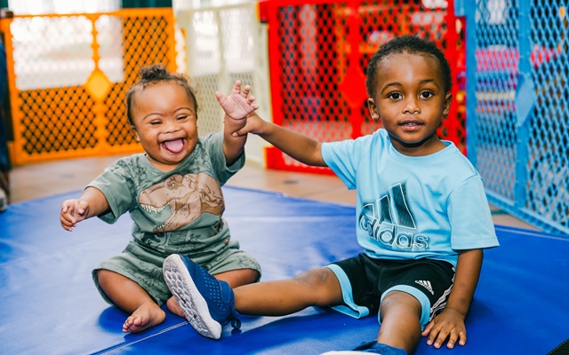 Two young boys on a blue mat, one laughing and the other smiling while holding hands