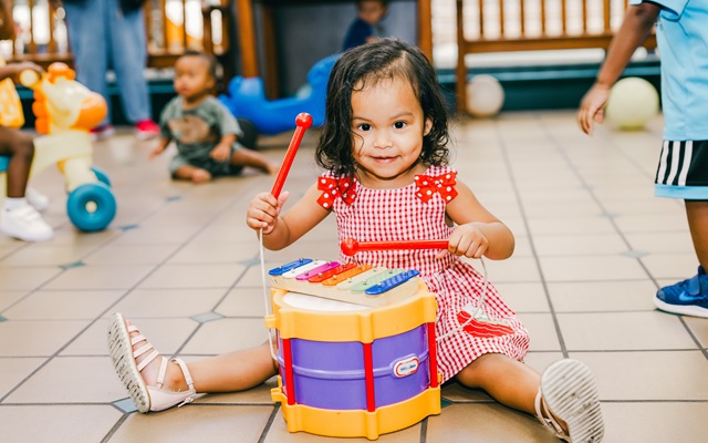 Little girl in red gingham dress happily playing a toy drum in a playroom