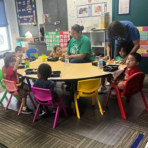 Children and adults sit around a classroom table on colorful chairs eating a snack