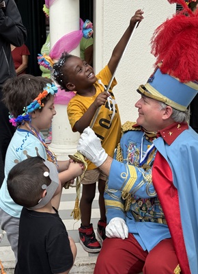 Children laughing and playing with a costumed King Antonio who wears a blue suit and red feathered hat