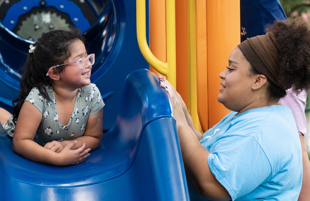 A girl wearing glasses smiles at her caregiver from a slide