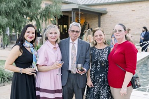 A group of five people in formal wear smile at the camera, a building and trees behind them