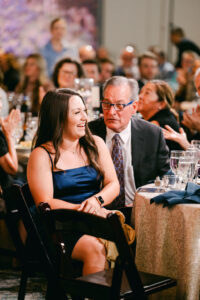 A lady in formal dress smiles at a table at a gala, many people behind her at other tables