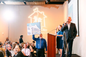 A male auctioneer on stage with a microphone, a man behind holds up a painting for sale, people in front sat at tables