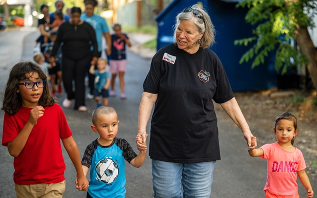 A smiling female caregiver holds the hands of children while walking outside, more children and caregivers in the background