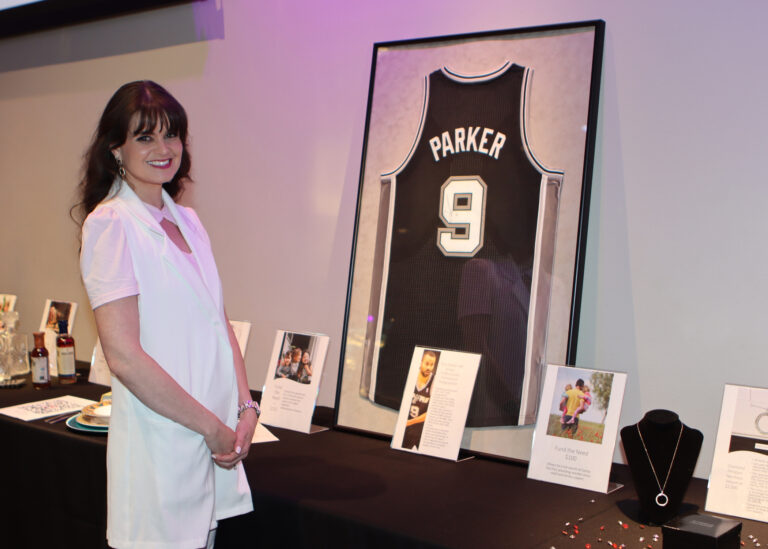 A girl in a white shirt standing in front of auction items, including a necklace and Spurs basketball jersey