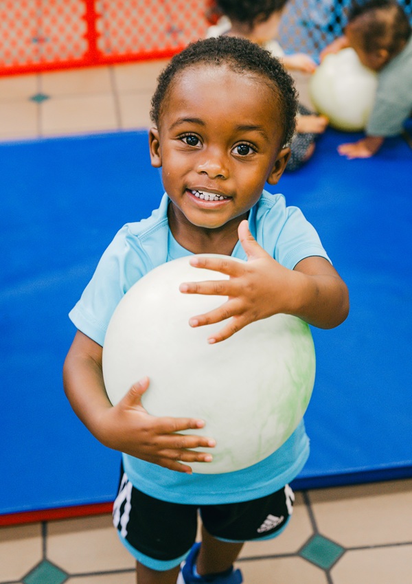 A smiling young boy in a blue shirt, looking at the camera clutching a white ball