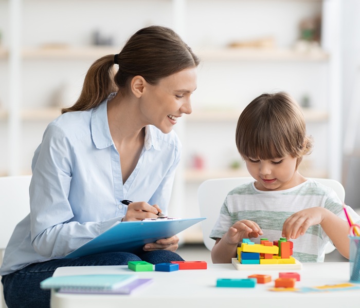 A woman smiling with a clipboard as a child plays with blocks in front of her at a table