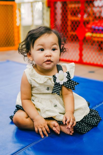Young toddler in a white dress with a flower and black polka dots sitting on a play mat