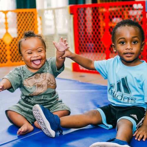 Two young boys on a blue mat, one laughing and the other smiling while holding hands