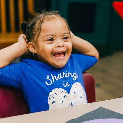Smiling young girl sits at a table wearing a blue shirt, hands behind her head.