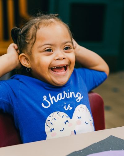 Smiling young girl sits at a table wearing a blue shirt, hands behind her head.