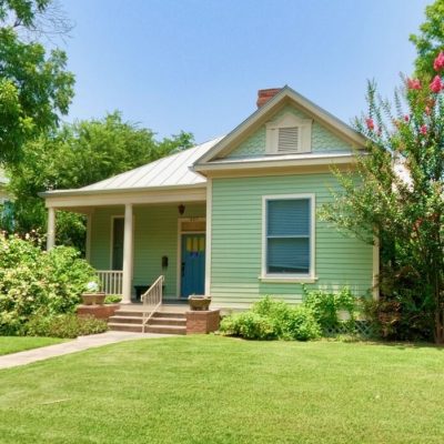 Light green house with a blue door and porch, surrounded by trees and flowers on a sunny day.