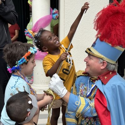Children laughing and playing with a costumed King Antonio who wears a blue suit and red feathered hat